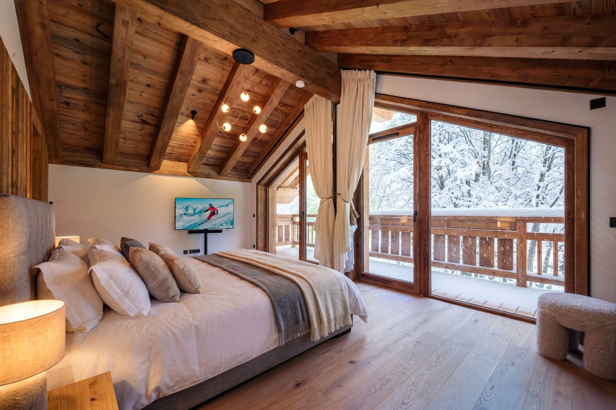 A cozy bedroom in a rustic alpine chalet with exposed wooden ceiling beams, a large bed with white linens and beige pillows, and floor-to-ceiling glass doors opening to a snowy outdoor balcony.