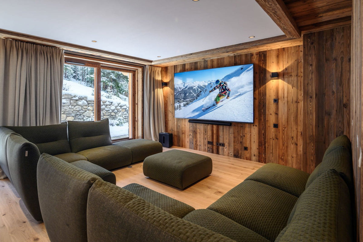A modern ski chalet living room featuring a large U-shaped olive green sectional sofa and matching ottoman, set against a wood-paneled wall with a large wall-mounted TV showing a skier on a snowy mountain.