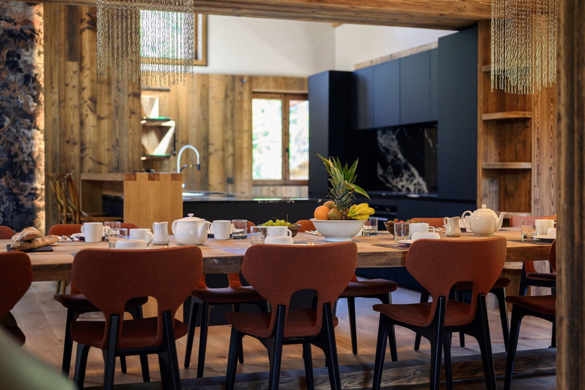 A rustic dining room scene with a long wooden table set with white teacups, a white teapot, and fruit, surrounded by reddish-brown upholstered chairs. The background features a modern kitchen with dark blue cabinets and wood-paneled walls.
