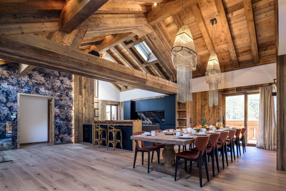 A luxury chalet-style dining room with a long wooden dining table and chairs, two large hanging crystal chandeliers, a stone wall, and a rustic wooden ceiling with exposed beams.