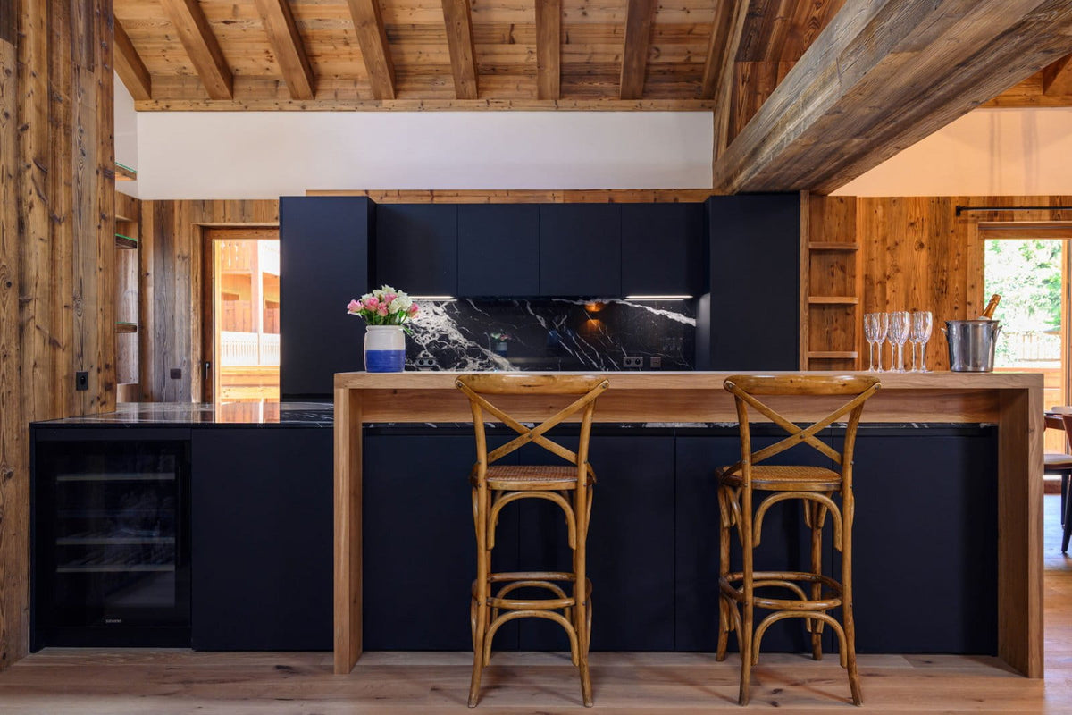 A modern kitchen island with matte black cabinetry, a light wood countertop, and two wooden bar stools in a rustic cabin interior with exposed wood ceiling beams.