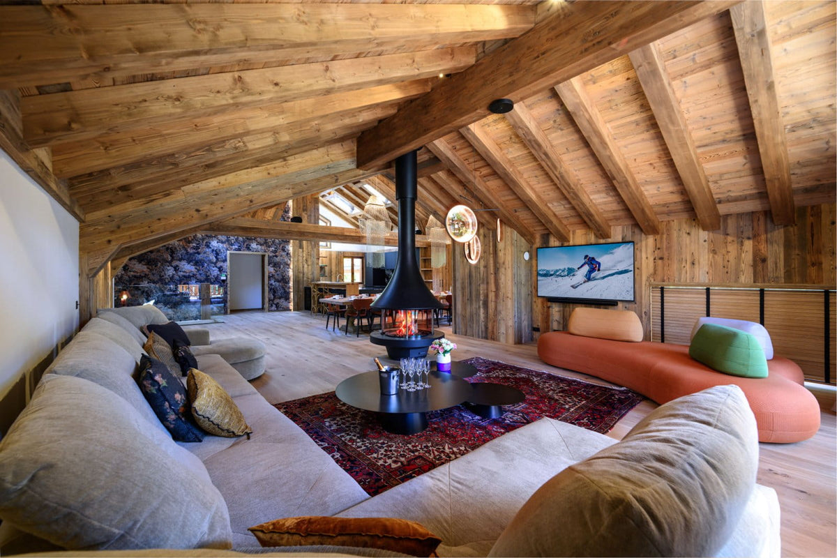 A cozy ski lodge living room with a large beige sectional sofa, a modern black fireplace, a red patterned rug, and a curved orange lounge chair under a rustic wooden ceiling.