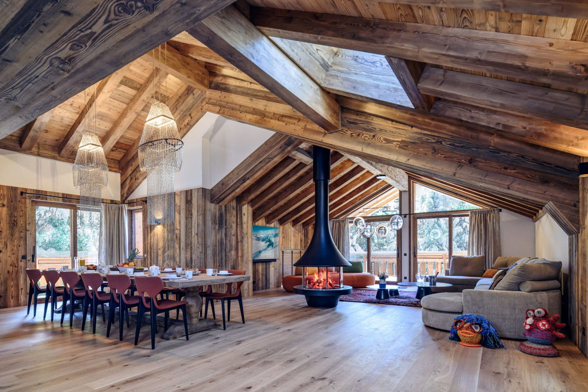 A high-ceilinged rustic lodge interior with exposed wooden beams, a central black flue fireplace, a dining table with chairs, a sectional sofa, and large windows looking out to a deck.