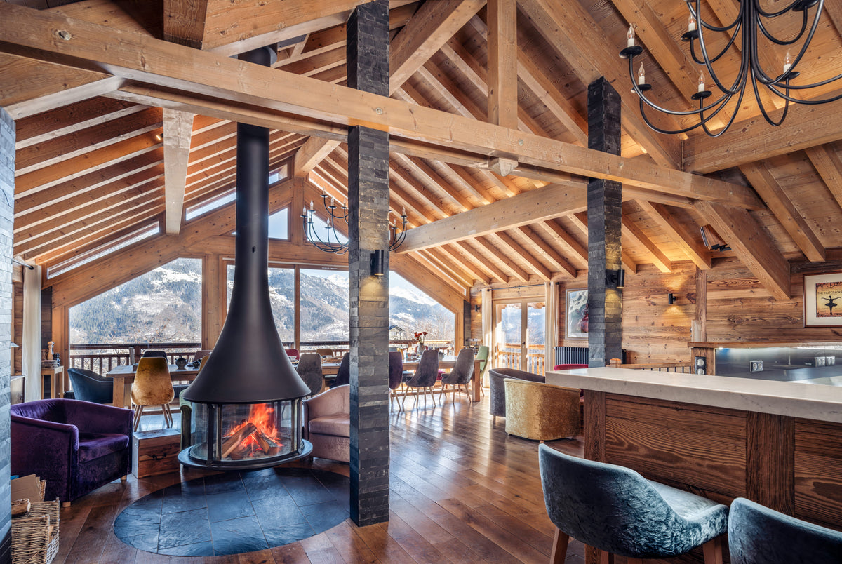 A large rustic lodge interior with exposed wood beams, a central black freestanding fireplace with visible flames, wood floors, bar stools, and mountain views through the windows.