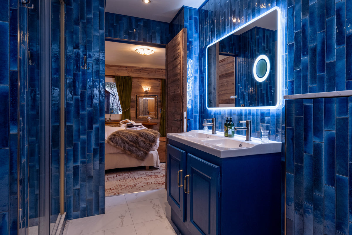 A luxurious modern bathroom featuring deep blue glossy tile walls, a matching blue vanity with a white sink countertop, an illuminated rectangular mirror, and a glass shower enclosure.
