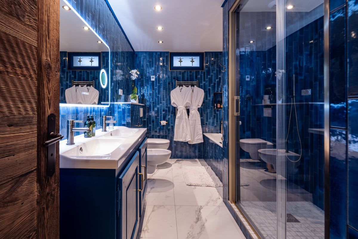 A luxury modern bathroom featuring deep blue tiled walls, a white marble floor, a double-sink vanity with a large mirror, a white robe hanging on a rack, and a glass-enclosed shower area.