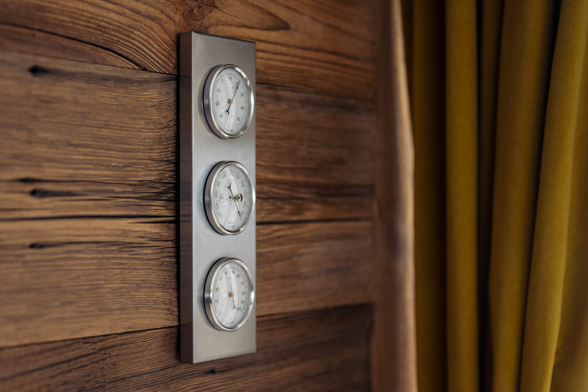 A vertical wall-mounted metal clock with three round analog dials, shown against a dark wood-paneled wall with a mustard yellow curtain on the right.