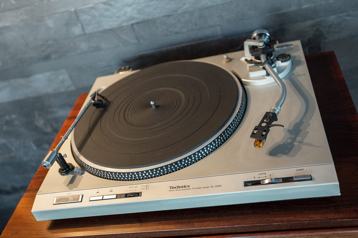 A Technics turntable with a beige outer shell and a dark gray platter, resting on a dark wood surface against a gray brick wall.