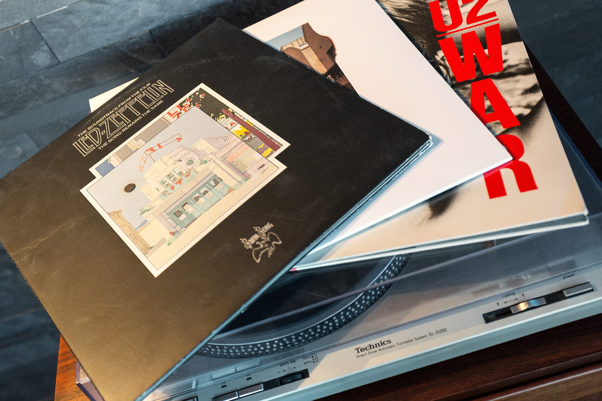 A stack of vinyl record albums resting on a Technics turntable. The top album cover is black with a small illustrated patch and the text 'Jazzitopia'; beneath it are white and cream covers with bold red lettering and a partial portrait.