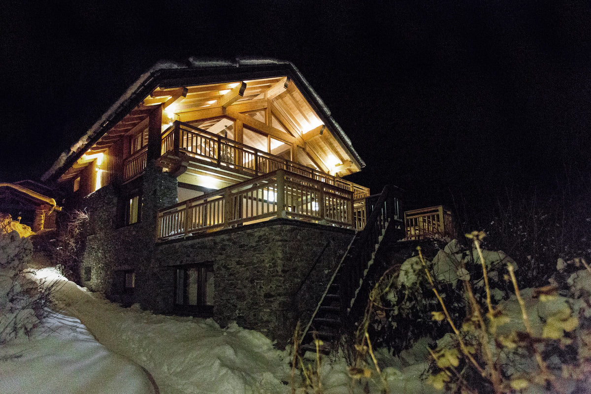 A snow-covered alpine chalet at night with warm yellow lights glowing from the windows and wooden balconies, surrounded by dark trees and snow on the ground.