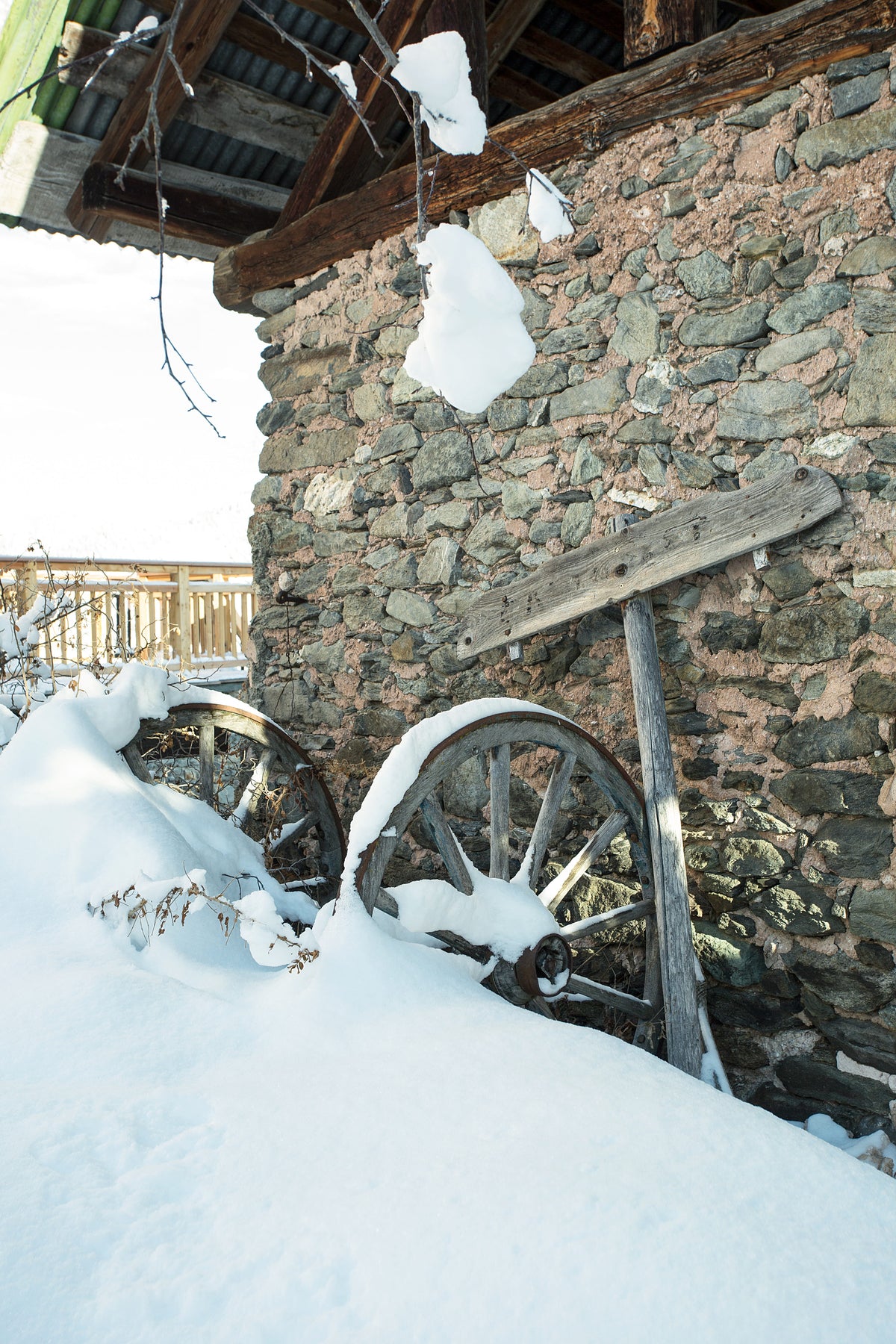A rustic stone wall beside a snow-covered wooden wagon wheel, with deep snow in the foreground and part of a building roof above.