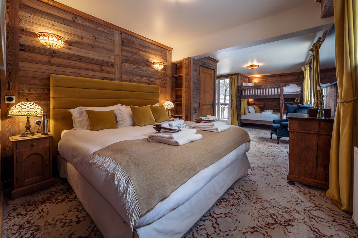 A rustic mountain-style bedroom featuring a large bed with a mustard yellow upholstered headboard, white bedding, a beige throw blanket, and matching accent pillows. The room has warm wood-paneled walls, bedside tables with lamps, a patterned carpet, and a glimpse into an adjacent loft-style bedroom in the background.