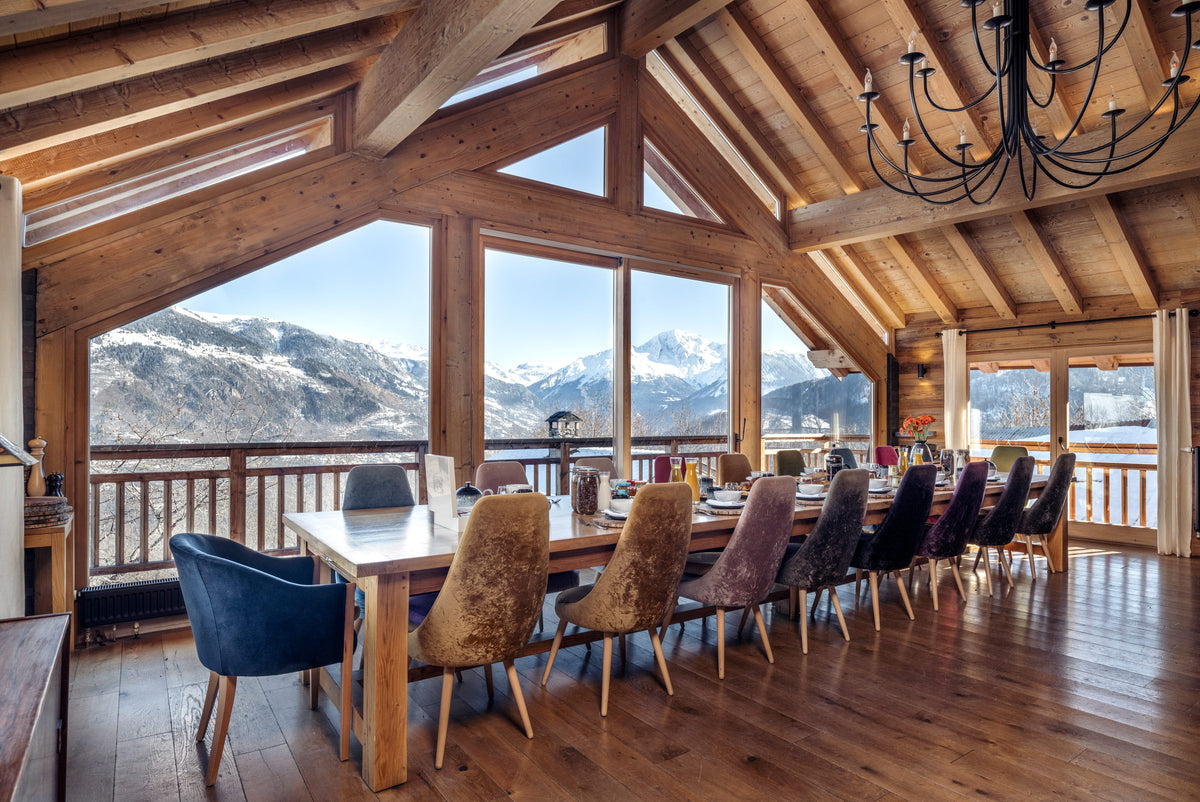A luxury alpine chalet dining room with a long wooden dining table and chairs, set under a high wood-beamed ceiling with a large chandelier, featuring panoramic windows overlooking a snowy mountain landscape.
