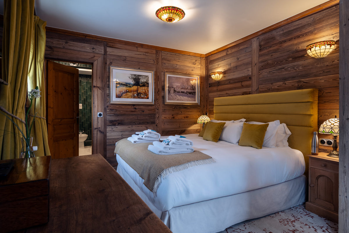 A rustic bedroom with wood-paneled walls, a large bed with white bedding and olive green pillows, a yellow upholstered headboard, and warm ambient lighting.