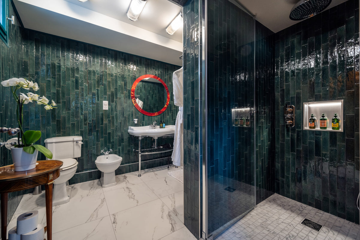 A modern bathroom interior with glossy dark green tiled walls, a glass shower enclosure, white marble floor, toilet, sink, and a red-rimmed oval mirror.