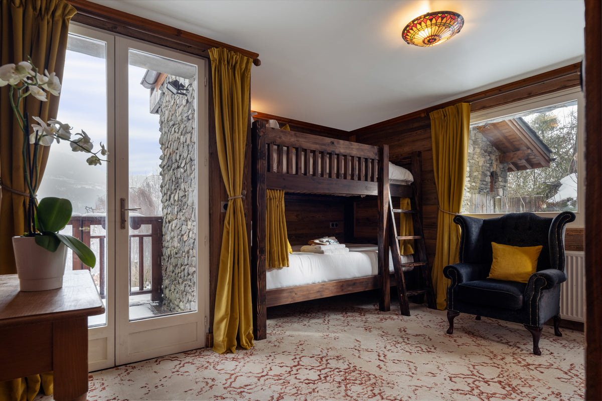 A rustic mountain chalet bedroom featuring a dark wood bunk bed, a black wingback armchair with a yellow pillow, mustard yellow curtains, and large windows looking out onto a snowy outdoor scene.