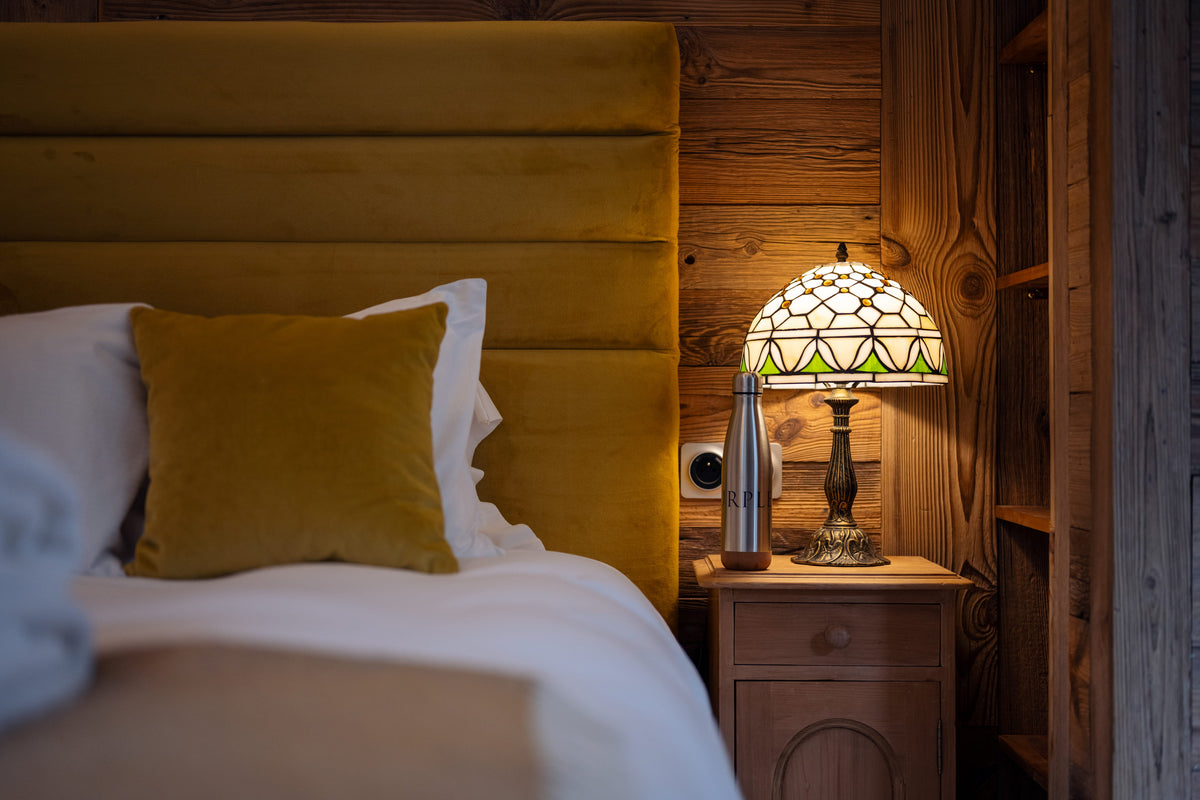 A cozy bedroom corner featuring a bed with white linens and a mustard yellow pillow in front of a padded yellow headboard, next to a wooden nightstand with a lit stained-glass table lamp and a metallic bottle.