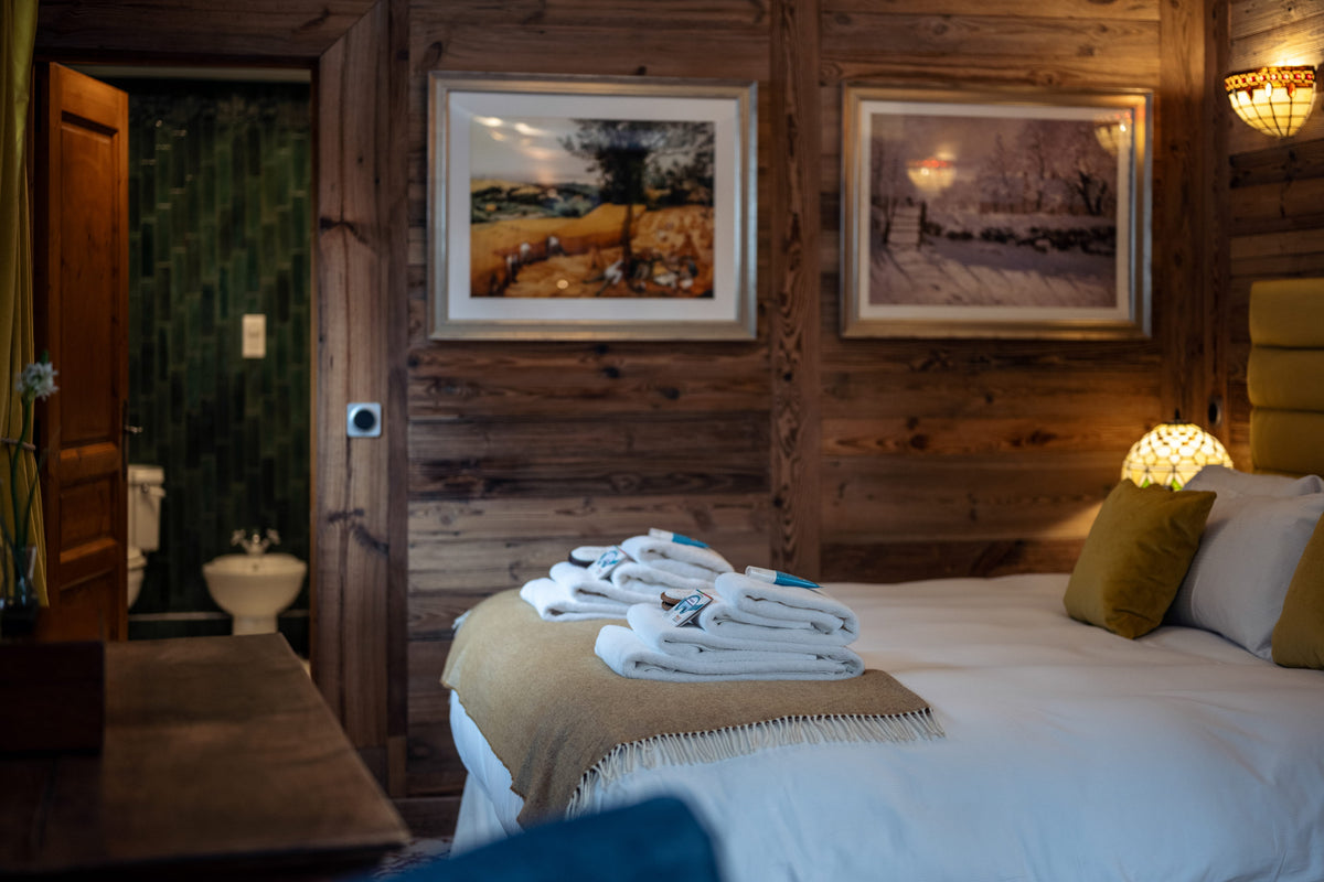 A cozy bedroom interior with a bed dressed in white linens and a beige throw, set against rustic wooden walls. Two framed landscape prints hang above the bed, and a stack of folded white towels sits on the foot of the bed.