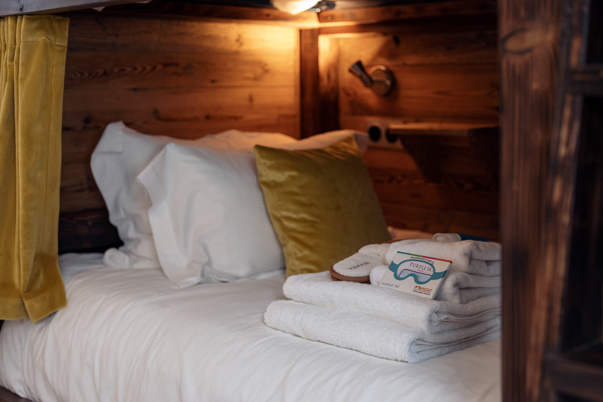 A cozy bed in a rustic wooden cabin with white bedding, white pillows, a gold accent pillow, and folded white towels on the bed.