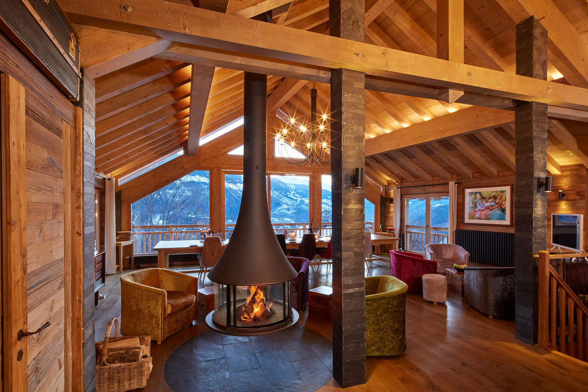 A rustic mountain chalet interior with a central black cone fireplace, exposed wood beams, stone pillars, and large windows overlooking snowy mountains.