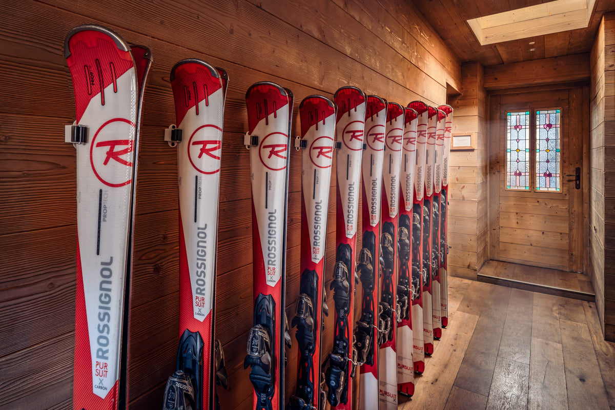 A row of Rossignol alpine skis with red and white graphics hangs on a wooden wall inside a rustic chalet with wood floors and a stained glass door.