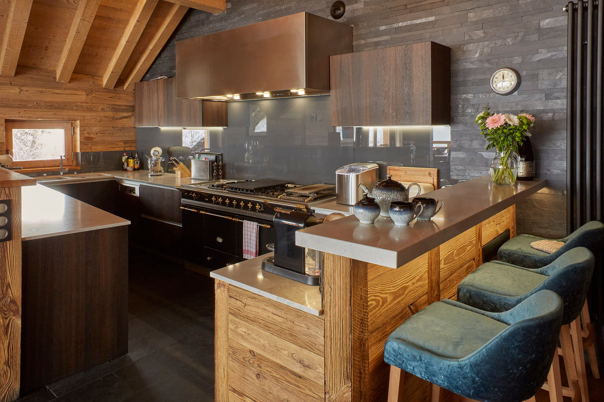 A modern alpine-style kitchen with dark wood cabinetry, a large stainless steel range hood, a gas stove, and a central island with a wood-look base and a dark countertop. Three blue upholstered bar stools sit at the island, and a vase of flowers and a round wall clock are visible on the counter.