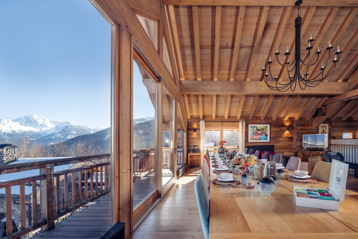 A bright, modern chalet dining room with a large wooden table set for a meal, overlooking a snowy mountain landscape on a sunny day. The room features a rustic wood ceiling, a black chandelier, and large windows.
