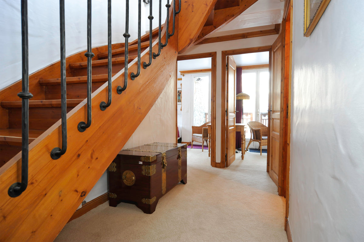 A home interior hallway featuring a prominent wooden staircase with black metal balusters on the left, a vintage brown trunk under the stairs, and an open doorway revealing a dining area in the background.
