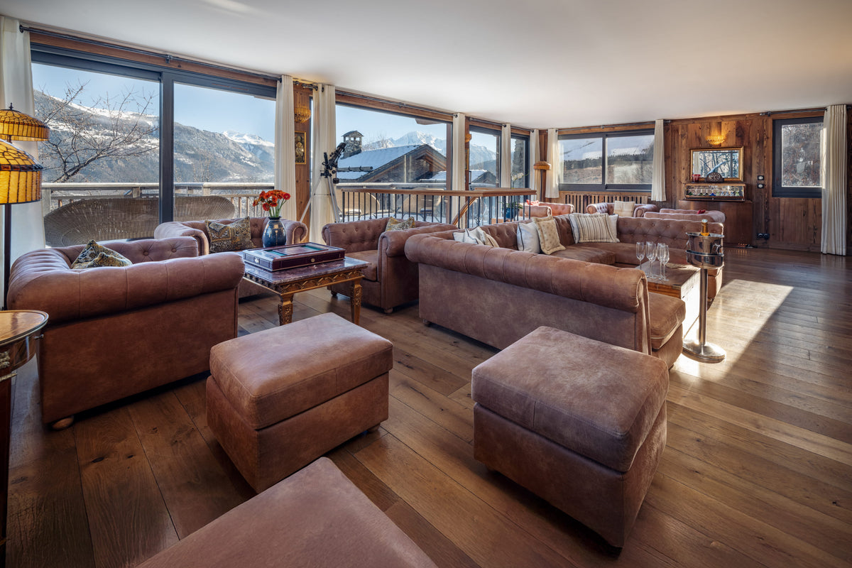 A luxury mountain chalet living room featuring plush brown leather sofas, matching ottomans, a wooden coffee table, and large floor-to-ceiling windows showcasing a scenic alpine view.
