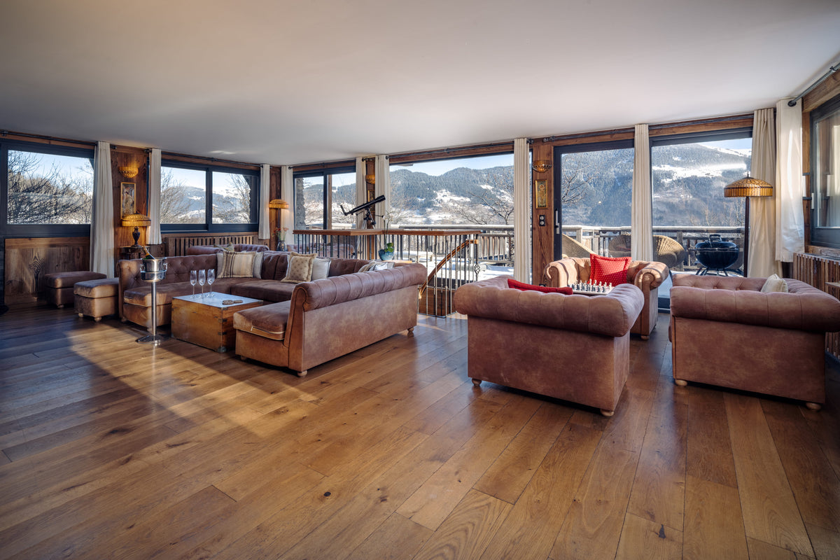 A spacious mountain-view living room with large windows, hardwood floors, and brown upholstered sofas and armchairs.