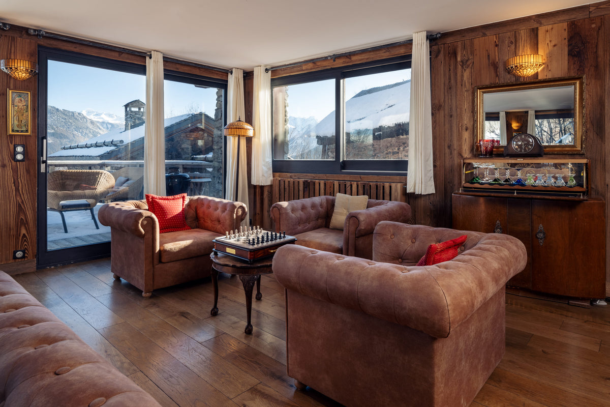 A cozy alpine chalet living room with brown leather armchairs, a small wooden coffee table, and large windows overlooking snowy mountains.