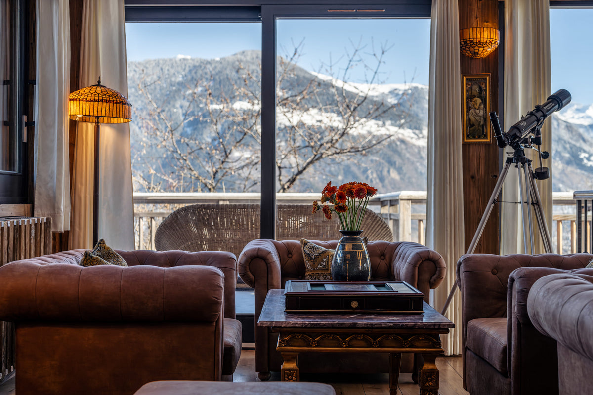 A luxurious mountain-view living room featuring brown leather armchairs, a dark wood coffee table with a vase of red flowers, a tripod-mounted telescope, and large floor-to-ceiling windows showcasing snow-capped peaks.