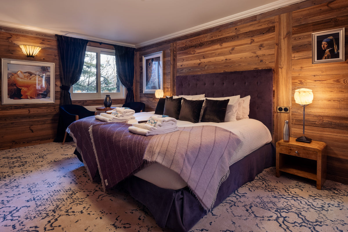 A cozy alpine-style bedroom with a large bed dressed in white and lavender bedding, set against rustic wood-paneled walls. A purple tufted headboard, patterned rug, bedside tables, and warm bedside lamps complete the scene.