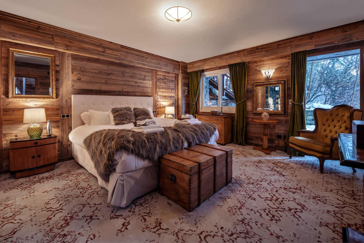 A rustic mountain-style bedroom with wood-paneled walls, a bed dressed in white linens and a brown fur throw, a wooden trunk at the foot of the bed, a patterned rug, and a tufted armchair by large windows.