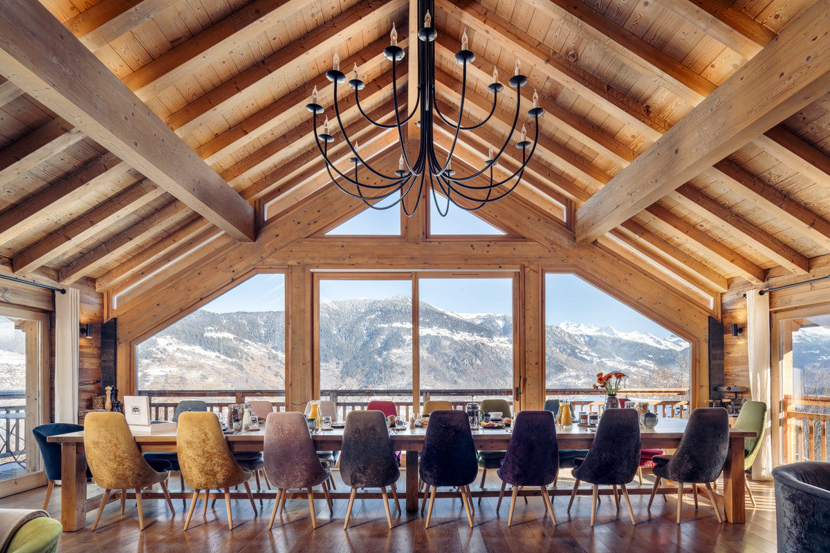 A luxury dining room with a long wooden table and ten upholstered chairs in various colors, set under a high wooden-beamed ceiling with a large black chandelier. The room features massive windows looking out onto a snowy mountain landscape.
