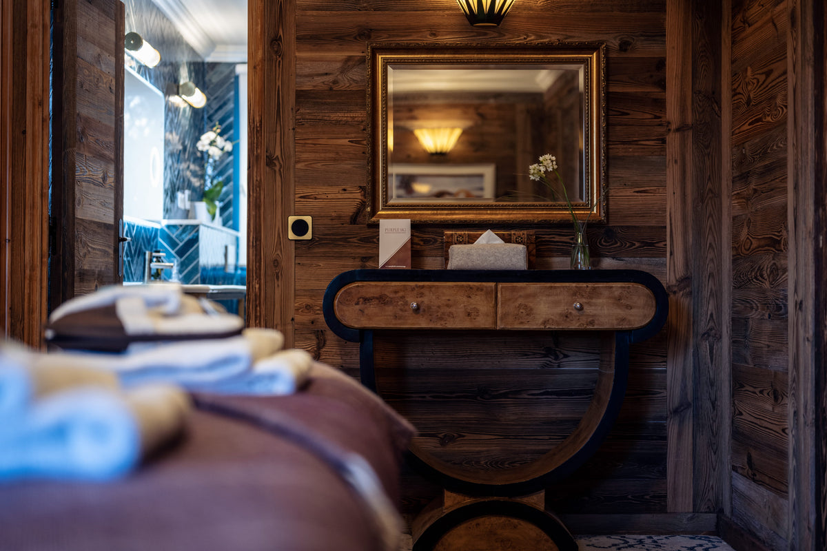A rustic wooden interior scene with a bed in the foreground, a wooden console table beneath a gold-framed mirror, and a doorway revealing blue-tiled walls and a bathtub in the background.