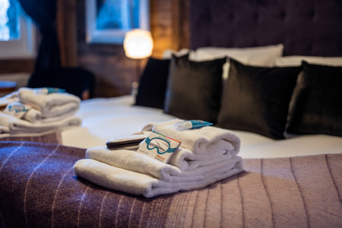 A stack of neatly folded white towels with a small blue and green tag rests on a bed in a cozy, dimly lit bedroom. The bed is made with white linens, black pillows, and a brown textured blanket.