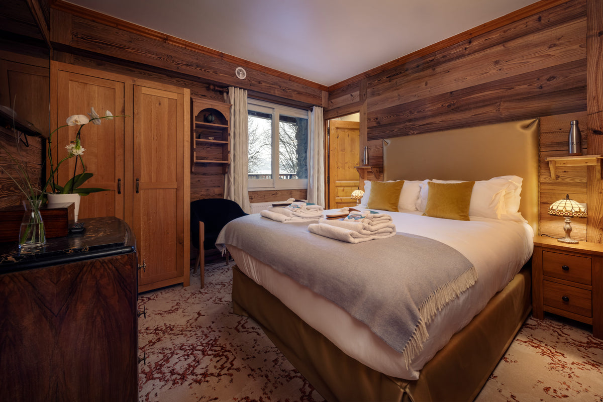 A cozy bedroom interior with a large bed dressed in white linens, a light gray throw blanket, and mustard yellow accent pillows. The room has warm wood-paneled walls, wooden furniture, a window, and bedside tables with lamps.