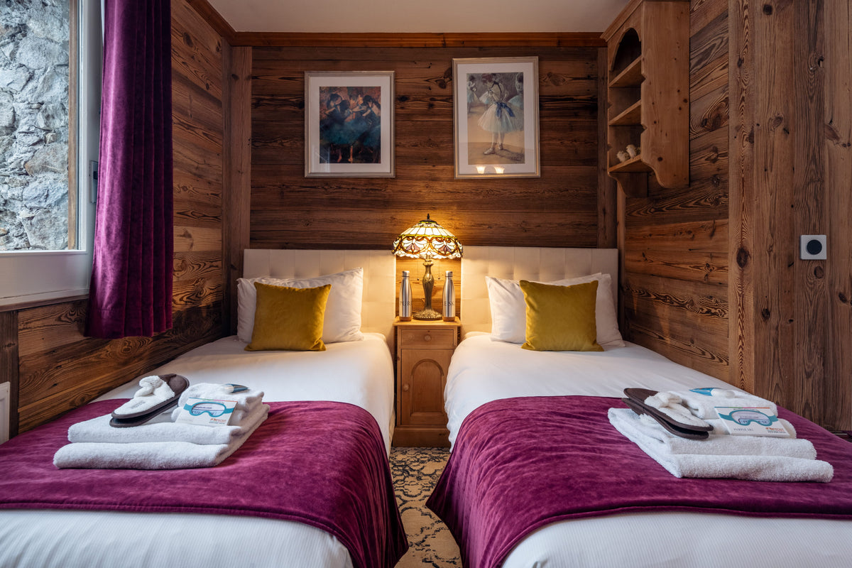 A cozy alpine-style bedroom with two single beds, wood-paneled walls, a lit bedside lamp, framed wall art, and purple bedding accents.