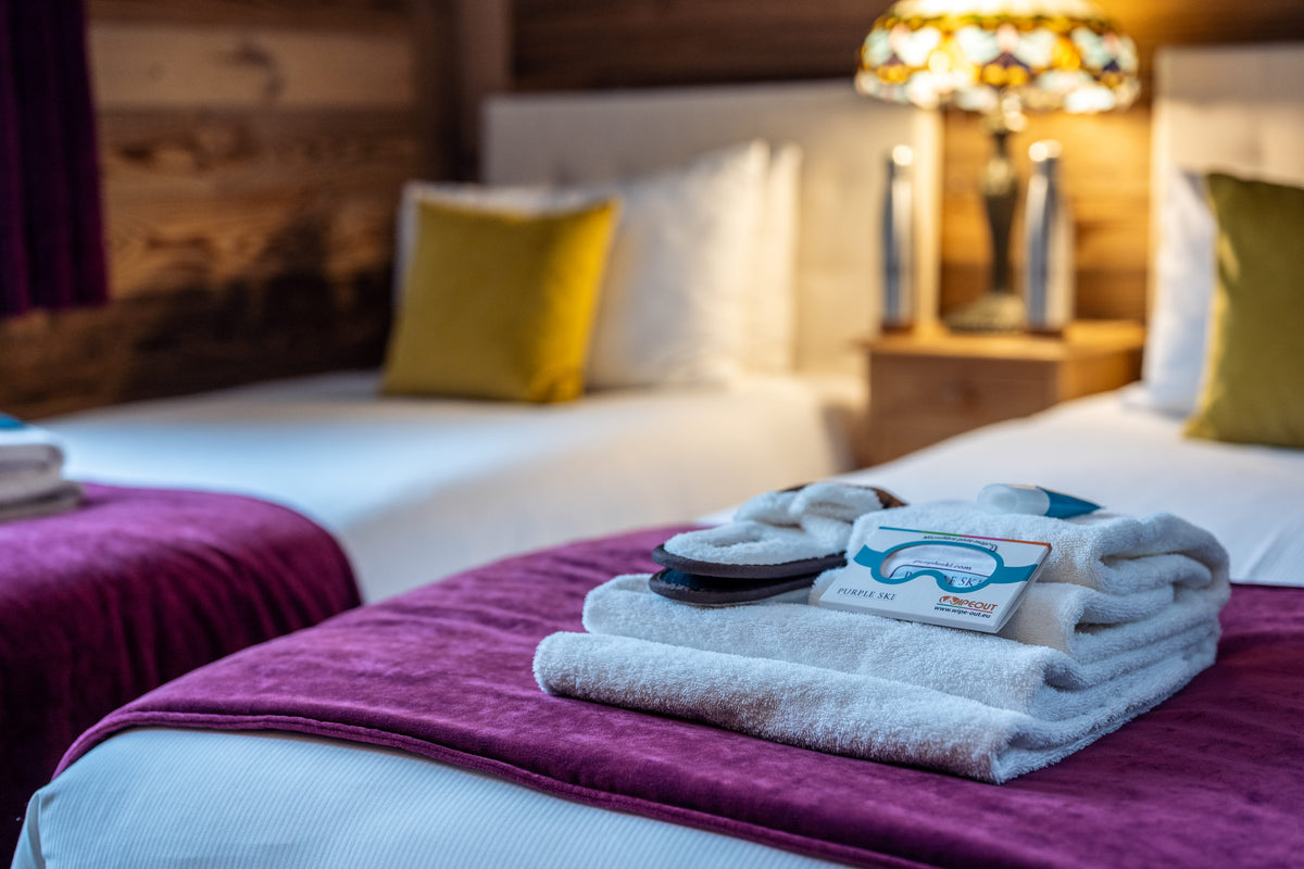 A set of white towels and slippers with a ski goggles tag, neatly folded on a bed with a purple blanket in a cozy, warmly lit bedroom.