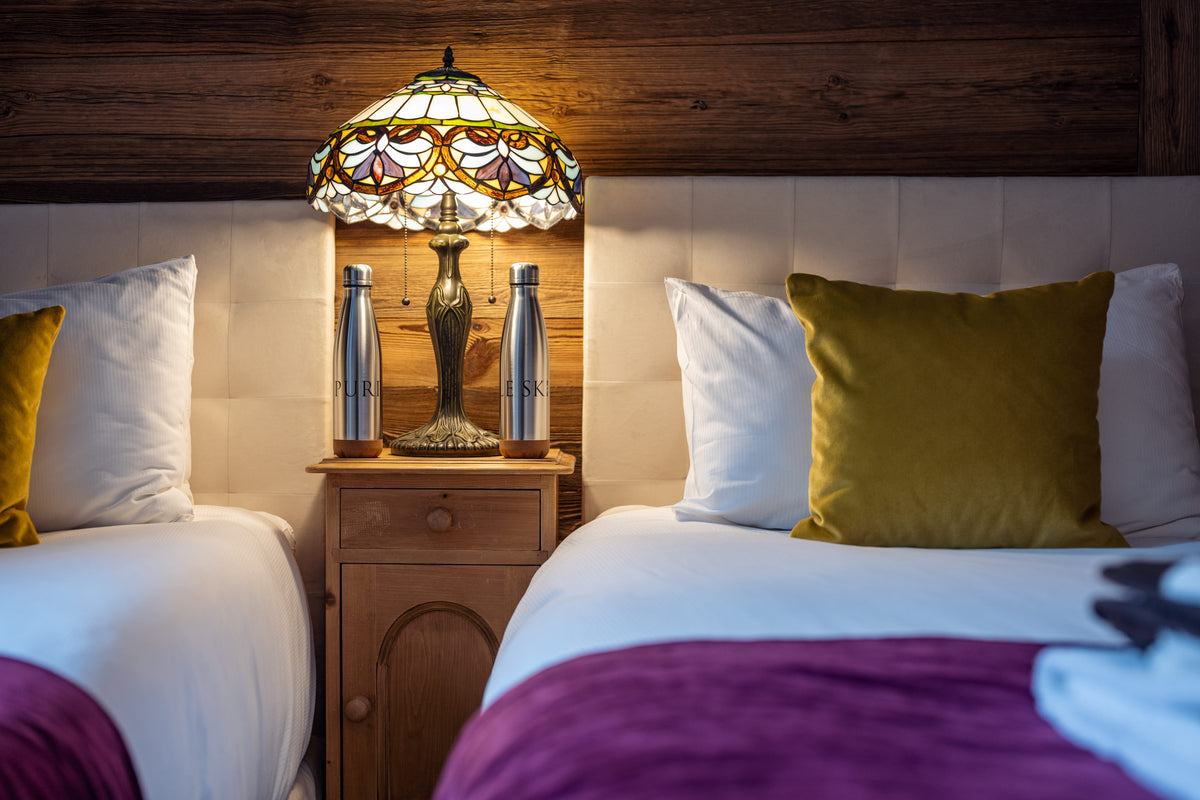 A warmly lit bedroom scene showing two neatly made beds with white linens, yellow accent pillows, and purple bedspread, centered around a wooden nightstand with a stained-glass table lamp and two metallic bottles.