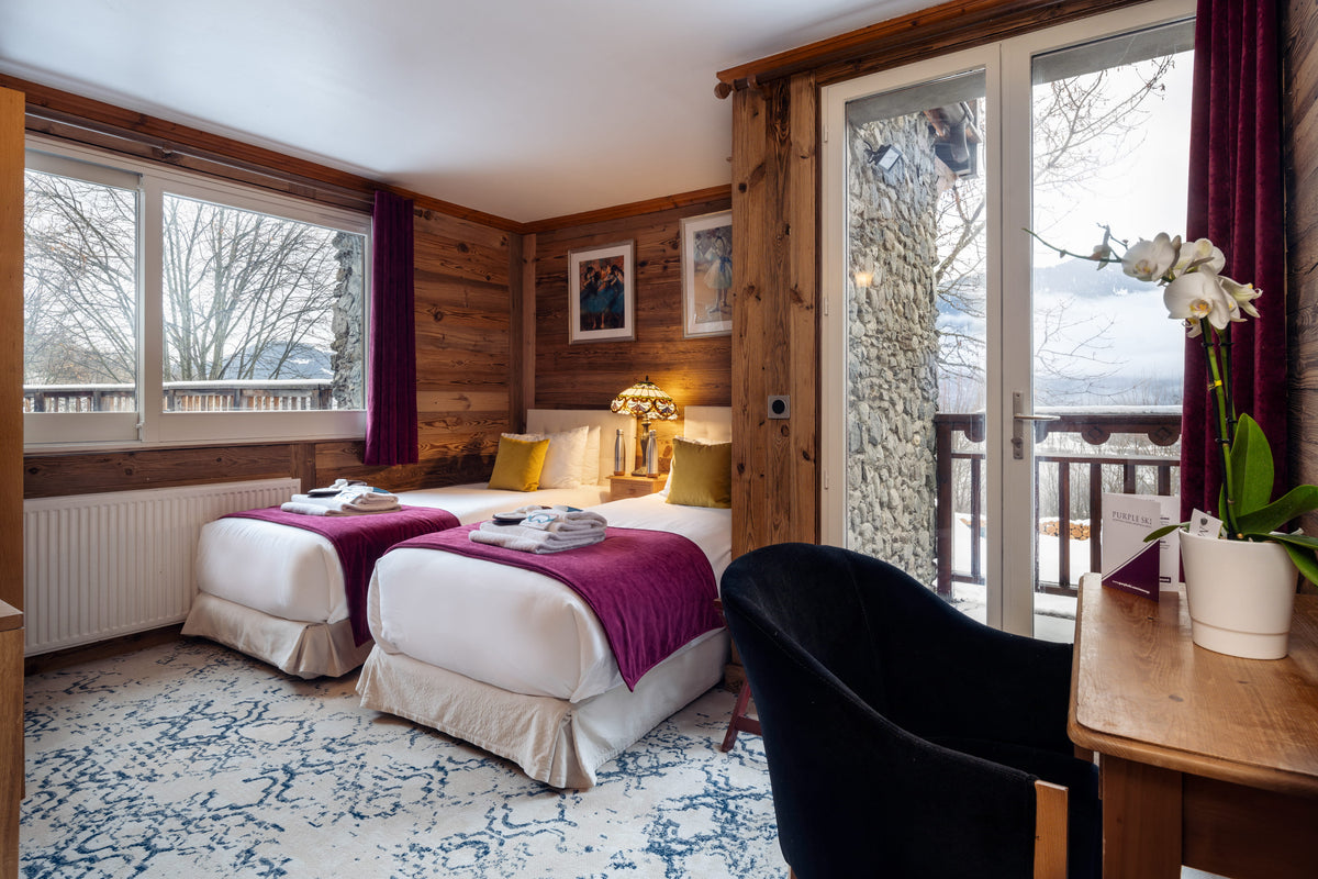A cozy ski chalet-style bedroom with two single beds dressed in white linens and deep purple throws, set against rustic wood-paneled walls. A large sliding glass door opens onto a balcony, and a black chair and wooden desk with a potted white orchid are in the foreground.
