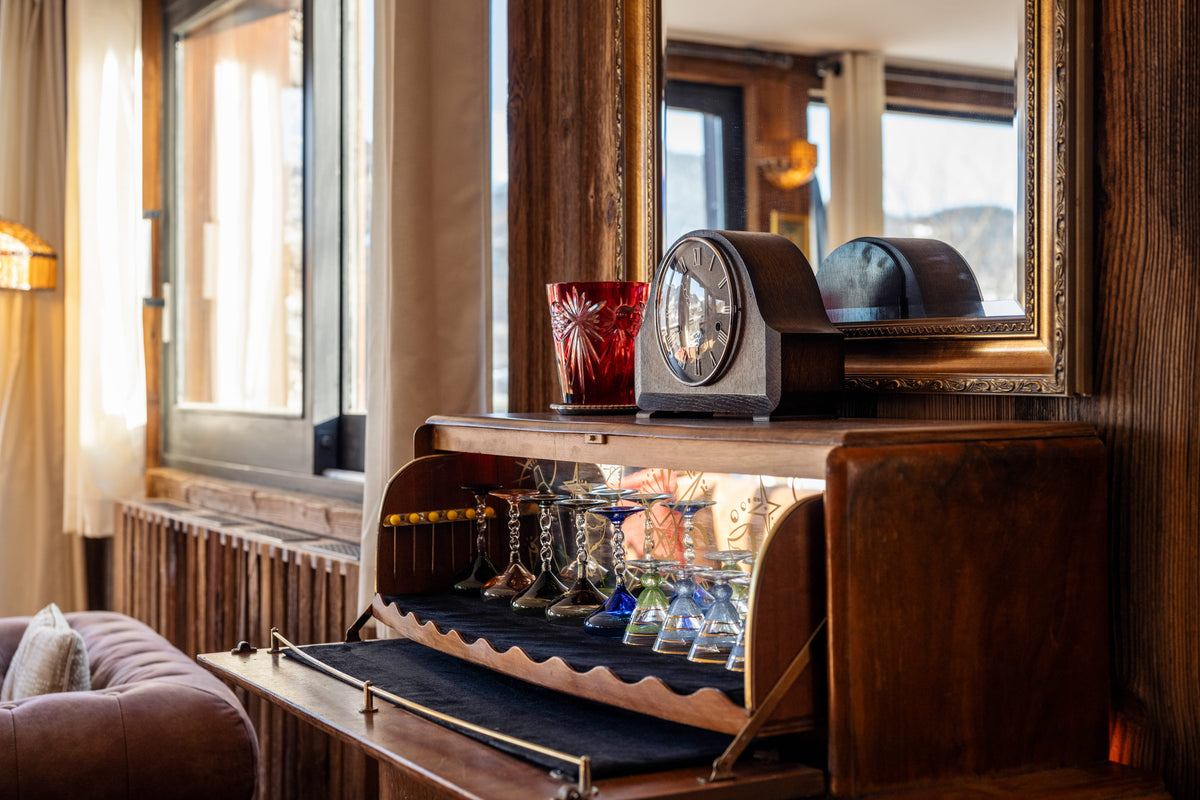 A vintage-style wooden bar cabinet with an open lower compartment displaying crystal glasses, set against a rustic wood-paneled wall with a large mirror and a red glass cup on top.