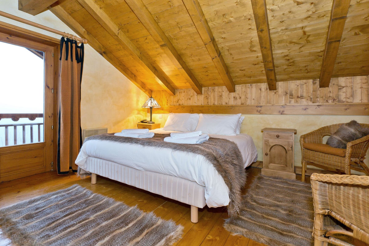 A cozy bedroom with a large bed dressed in white linens and a brown fur throw, set in a rustic wooden room with a slanted ceiling, wicker chair, and striped rugs.