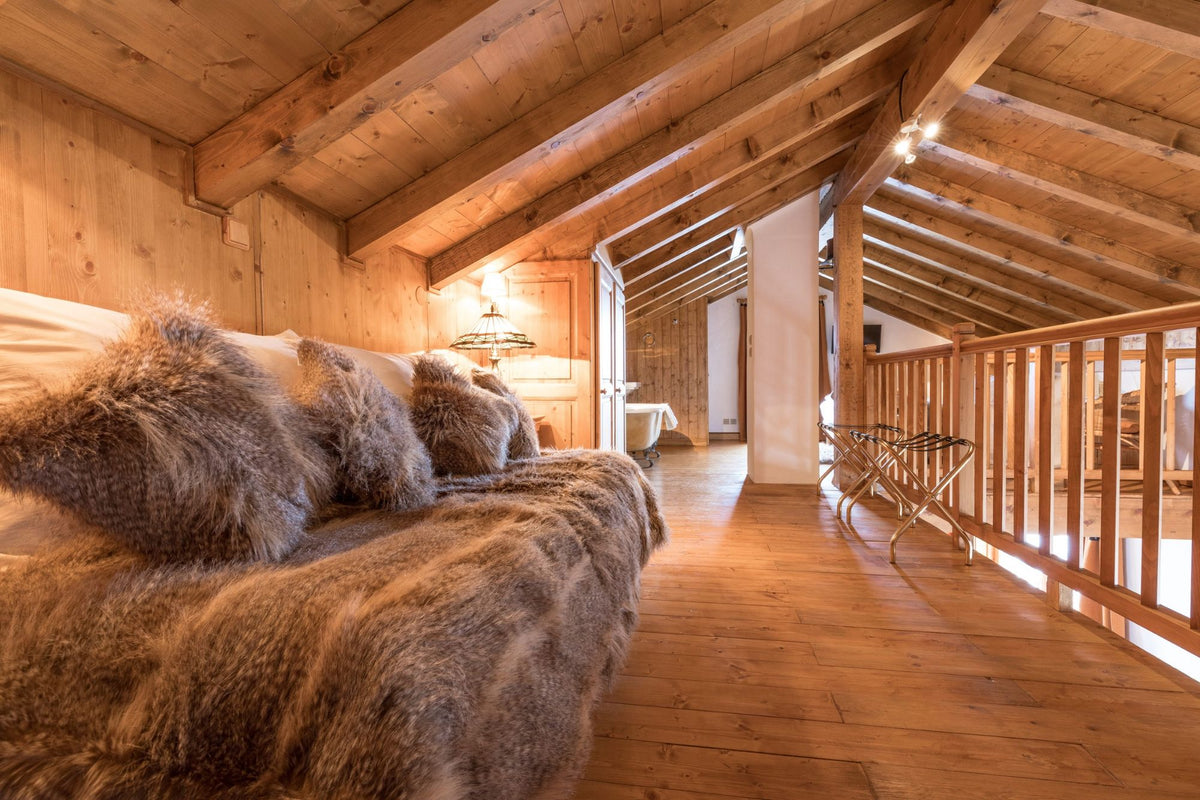 A cozy bedroom interior with a bed dressed in shaggy brown faux fur bedding and matching pillows, set in a rustic wooden chalet with exposed wood beams, a wooden staircase railing, and warm ambient lighting.