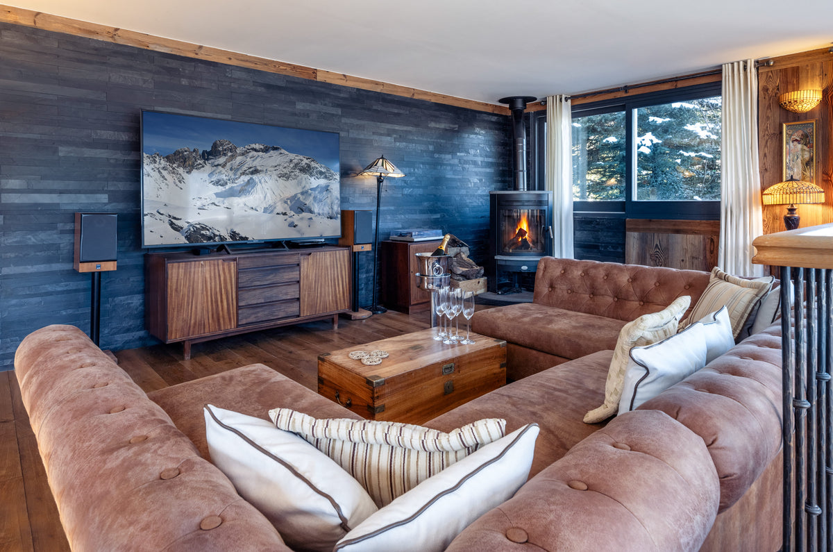 A cozy ski chalet living room featuring a large tufted brown sectional sofa with cream and striped pillows, a wooden coffee table, a dark wood media console with a large landscape TV, and a wood-burning fireplace.