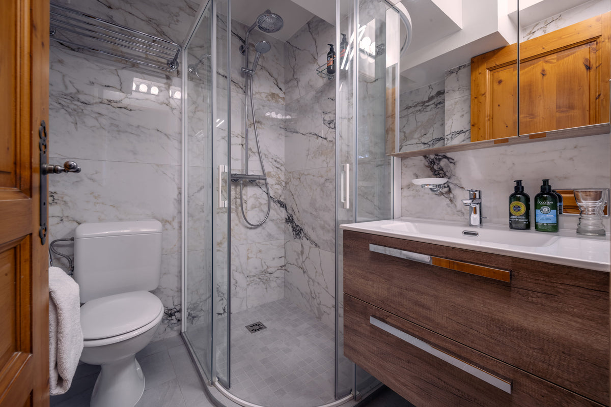 A modern bathroom interior featuring a glass shower enclosure, white marble walls and floor, a white toilet, a wood vanity with a white sink, and wooden wall cabinets.