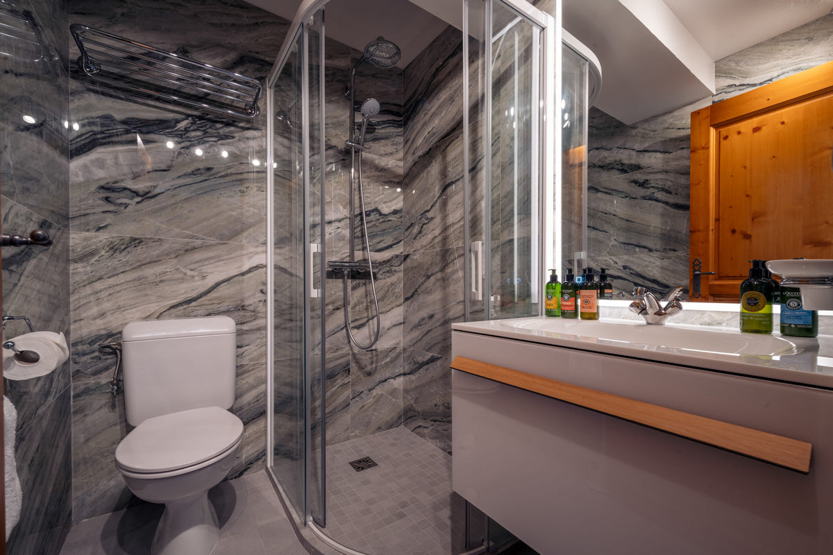 A modern bathroom interior featuring a glass shower enclosure, a white toilet, a floating vanity with a white sink, and gray marbled walls. A wooden cabinet is visible on the right, and small toiletry bottles sit on the counter.