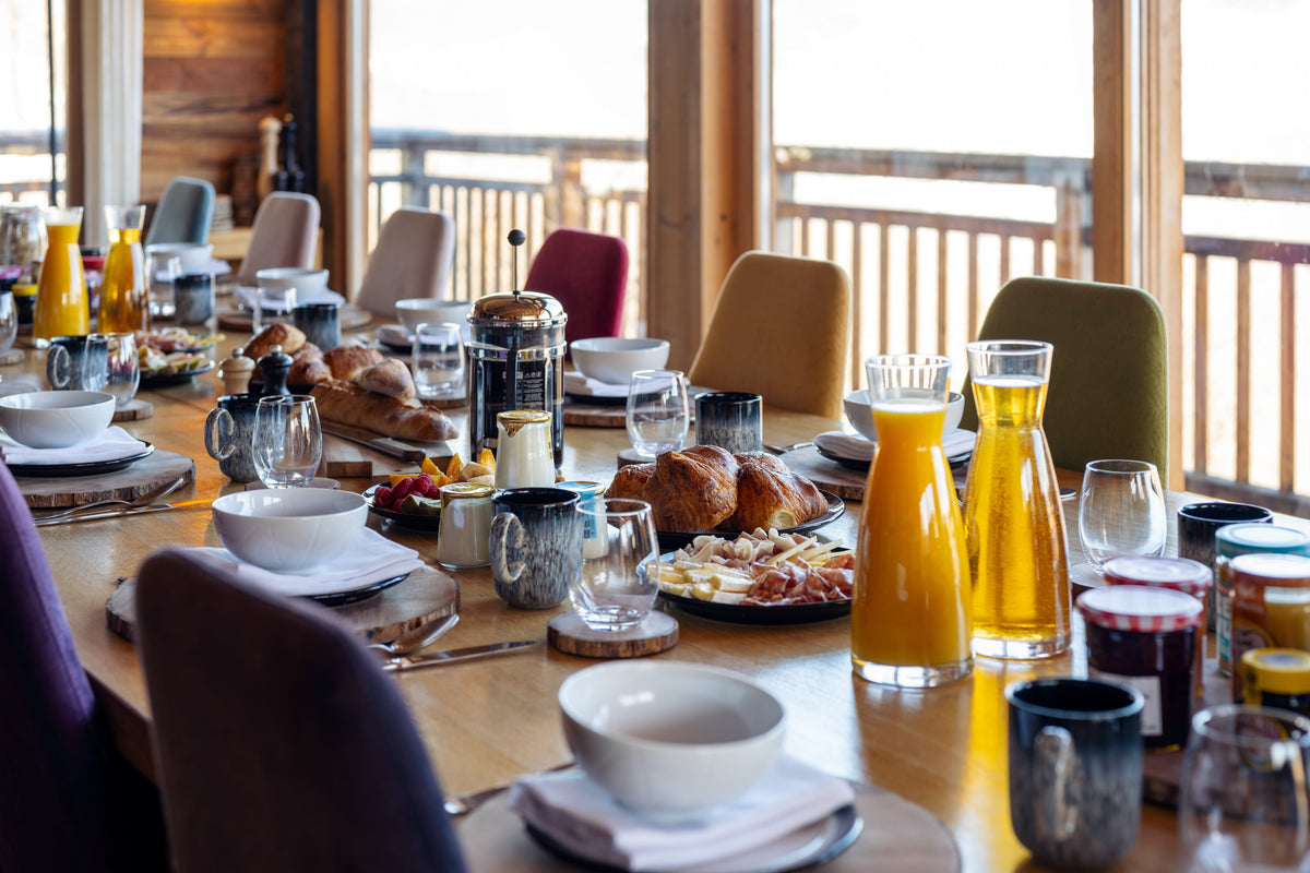 A rustic dining table set for breakfast in a cabin, featuring white bowls, plates, bread, fruit, and glass carafes filled with orange juice and coffee, with wooden chairs and large windows in the background.