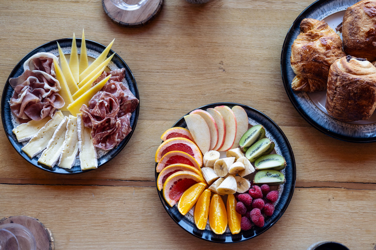 Three black plates arranged on a wooden table, featuring a charcuterie board with sliced cheeses, cured meats, and yellow fruit, a fruit platter with sliced apples, oranges, kiwi, raspberries, and bananas, and a plate of pastries.